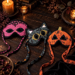 May include: Three fabric eye masks with long ties. The left mask is pink with black patterns, the center mask is dark gray with a cat-like shape and yellow eye accents, and the right mask is orange with a pumpkin-inspired design and black details.