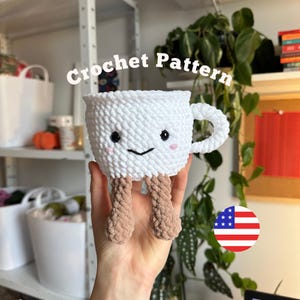 Könnte beinhalten: Eine weiße gehäkelte Tasse mit Henkel, schwarzen gestickten Augen, einem kleinen schwarzen Lächeln und rosa Wangen. Die Tasse hat zwei baumelnde, hellbraun gehäkelte Beine mit Füßen. Der Text Crochet Pattern ist über der Tasse sichtbar.