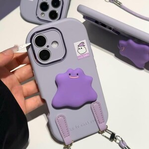 May include: A light purple smartphone case featuring a raised purple blob character with a simple smile, a small sticker of a ghost wearing a hat with the text "nice baby", and a matching purple strap attached with silver metal hardware.