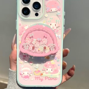 May include: A clear phone case with a light mint green border featuring pink My Melody character graphics, small floral accents, and the text My Piano. A circular pink shaker attachment filled with small loose pink charms is centered on the back.