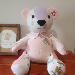 May include: A patchwork teddy bear constructed from various light pink, white, and lavender fabric panels. The bear features a black nose, dark button eyes, a white collar with lace trim, and embroidered cartoon character graphics on its limbs.