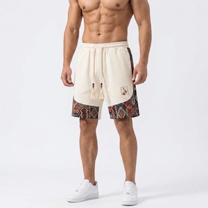 Peut inclure: Shorts blanc cassé avec une taille à cordon de serrage ornée de perles en bois et de pompons. Les panneaux inférieurs présentent un motif géométrique multicolore dans des tons orange, bleu et marron. Un petit écusson carré est visible.