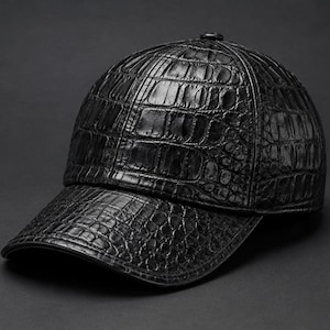 May include: A black baseball cap featuring a prominent, glossy, textured crocodile skin pattern across the entire surface, including the rounded crown and the curved brim, with a small button detail at the very top centre of the cap.