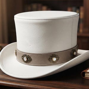 May include: A white top hat featuring a flat crown and a wide, curved brim. A taupe-colored band encircles the base of the crown, decorated with a series of evenly spaced, ornate metallic filigree accents, each centered with a small white pearl.