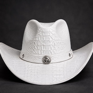 Puede incluir: Un sombrero de vaquero blanco con un patrón de textura de piel de cocodrilo en la copa y el ala. El sombrero incluye una banda blanca a juego adornada con tachuelas de metal plateado y un emblema circular central con un diseño de cabeza de toro.