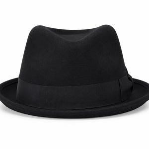 Może przedstawiać: Jednolity czarny kapelusz typu fedora ze strukturalną koroną z centralnym zagięciem i zwężanym przodem. Kapelusz posiada szeroką, pasującą czarną wstążkę rypsową otaczającą podstawę korony, wykończoną małą, płaską kokardką.