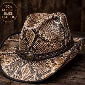 May include: A cowboy hat featuring a textured snakeskin pattern in shades of tan, brown, and black. The hat includes a dark brown braided leather band accented with a central metallic star emblem. Text on the image reads 100% GENUINE SNAKE LEATHER.