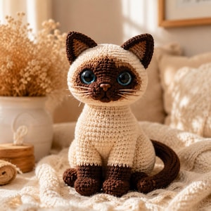 May include: A crocheted Siamese cat soft toy featuring a cream-coloured body with dark brown ears, face, paws, and tail. The cat has large, round, glossy blue eyes, a small brown nose, and stitched whiskers, sitting in an upright position.