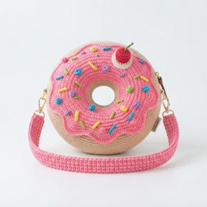 Pode incluir: Uma bolsa tiracolo redonda de crochê em formato de donut com base bege e cobertura rosa. A bolsa apresenta granulados de lã coloridos e texturizados em azul, amarelo, verde e roxo, com uma pequena cereja vermelha no topo e alça rosa.