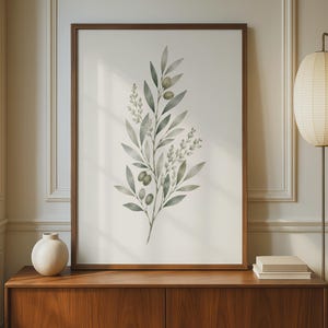 May include: A framed wall art print featuring a watercolor illustration of an olive branch with green leaves and small olives. The artwork is displayed in a dark wood frame, centered on a wooden sideboard next to a round vase and a stack of books.