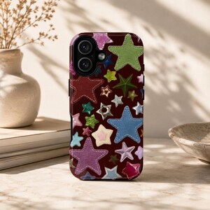 Op de afbeelding: Een donkerbordeauxrode smartphonehoes met een herhalend patroon van veelkleurige sterren in verschillende formaten. De sterren zijn uitgevoerd in tinten groen, blauw, roze, paars en wit, met zichtbare stikseldetails langs de randen van elke ster.