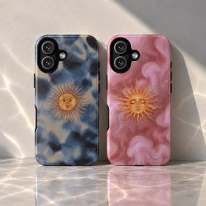 May include: Two rectangular phone cases featuring a sun motif. The left case has a dark blue and black tie-dye pattern with a gold sun. The right case has a pink and white cloudy marble pattern with a gold sun. Both have black camera cutouts.