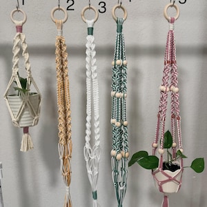 May include: Five macrame plant hangers numbered one through five, suspended from a metal rod by wooden rings. The hangers feature various knot patterns, colors including cream, yellow, green, and pink, and some incorporate wooden beads.