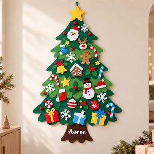 May include: A green felt Christmas tree wall hanging featuring a yellow star topper and various detachable ornaments including a Santa face, snowman, gingerbread man, candy canes, snowflakes, and gift boxes. The brown trunk displays the name Aaron.
