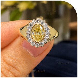 May include: A gold ring featuring a central oval-cut yellow gemstone surrounded by a halo of small, round, clear white stones. The band is smooth and polished gold, with the setting creating a scalloped edge around the central yellow stone.