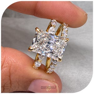 Puede incluir: Un anillo de oro con una gran piedra central rectangular de corte radiante sujeta por cuatro garras. La banda está dividida en dos secciones, cada una acentuada con piedras más pequeñas redondas y en forma de pera, con acabado pulido.