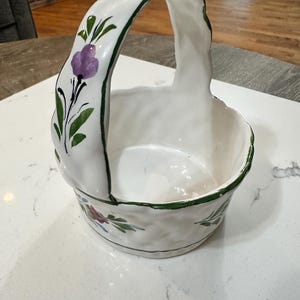 May include: A white ceramic basket with a high arched handle, featuring hand-painted green trim along the edges and floral motifs in purple, red, and green. The basket has a textured, wavy surface and a round base, showcasing a glossy finish.