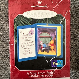 Può includere: Una scatola di ornamento Hallmark Keepsake con una scena di Winnie the Pooh. La scatola mostra un ornamento blu a forma di libro con Pooh e Pimpi all'interno. Il testo sulla scatola recita: Thanks for the honey, said Pooh. Is there more?