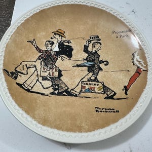 May include: A round ceramic decorative plate with a tan background featuring a Norman Rockwell illustration titled Promenade à Paris. The image depicts two men walking, one carrying a Vote for Al sign and the other a suitcase labeled Egbert.