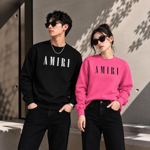 May include: A black long-sleeve sweatshirt with the white text AMIRI on the chest and a bright pink long-sleeve sweatshirt with the black text AMIRI on the chest. Both garments feature a crew neckline and long sleeves with ribbed cuffs.