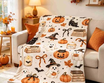 May include: A cream-colored throw blanket featuring a repeating Halloween pattern of orange pumpkins, white ghosts, black cats, brown autumn leaves, black bows, candles, and stacks of books with text including "Spooky Season" and "Tricks or Treat".