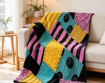 May include: A plush throw blanket featuring a patchwork design with alternating sections of yellow, teal, and pink. The pattern includes black stripes, polka dots, swirls, and grid lines, all separated by thick black borders in a quilt-like style.