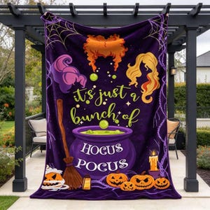 May include: A purple fleece blanket featuring a graphic print of three distinct hairstyles, a broomstick, a bubbling cauldron, and jack-o'-lanterns. The text reads, "it's just a bunch of HOCUS POCUS" in yellow and white lettering against a dark background.