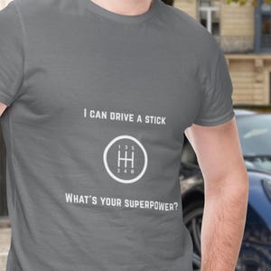 May include: A grey short-sleeved t-shirt featuring white text that reads I CAN DRIVE A STICK and WHAT'S YOUR SUPERPOWER? with a white graphic of a manual transmission gear shift pattern showing positions 1, 3, 5, 2, 4, and R inside a circle.