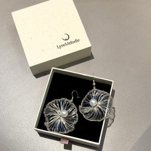 Puede incluir: Un par de pendientes de hilo plateado con un diseño circular tipo red y una perla blanca central. Los pendientes se presentan en una caja abierta de color crema con interior negro, que muestra el texto LyreMelodie y un logotipo de media luna.