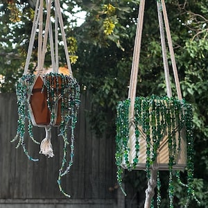 Op de afbeelding: Twee macramé plantenhangers gemaakt van off-white koord, elk met een pot. Eén pot is van terracotta, de andere is wit. Beide zijn versierd met lange, trapsgewijze strengen van gefacetteerde groene en blauwe glaskralen die ranken nabootsen.