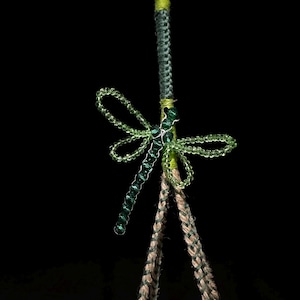 Può includere: Un ornamento a forma di libellula realizzato con perline verdi traslucide, caratterizzato da un lungo corpo segmentato e quattro ali delicate. La libellula è fissata a un cordino verticale avvolto da filo verde lime e grigio scuro, che si divide in due cordini intrecciati color marrone chiaro.