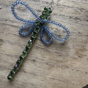 Può includere: Un ornamento a forma di libellula realizzato con filo metallico e perline sfaccettate. Il corpo è composto da perline verde scuro, mentre le quattro ali sono delineate da perline azzurre. Un piccolo occhiello in filo è fissato in cima.