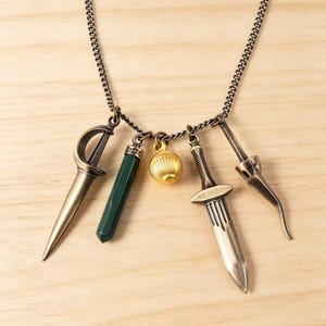 Peut inclure: Un collier composé d'une fine chaîne en métal sombre avec quatre breloques suspendues : une épée en bronze à garde incurvée, une pierre cylindrique vert foncé, une petite sphère dorée striée et une dague en bronze à pointe effilée.