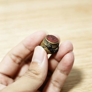 May include: A small, antique-style brass seal featuring a rounded body with intricate engraved patterns and a dark, weathered finish. The top of the seal is capped with a circular, flat red wax insert that displays a detailed embossed design.