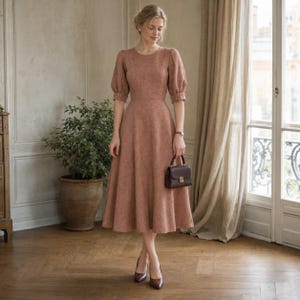 Puede incluir: Un vestido midi de color rosa polvoriento con cuello redondo, mangas abullonadas y una cintura entallada que se ensancha en una falda amplia. El vestido presenta una tela texturizada y se combina con un bolso estructurado marrón oscuro y tacones.