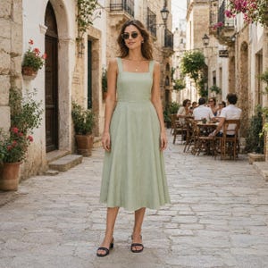 May include: A sage green midi dress featuring a square neckline, thick shoulder straps, and a fitted bodice that transitions into a flared, A-line skirt. The dress is made of a textured fabric and is paired with black strappy sandals and sunglasses.