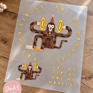 Könnte beinhalten: Ein durchscheinendes Blatt mit zwei Illustrationen eines braunen Affen, der einen gestreiften Partyhut trägt und goldene Becken hält. Der Text "MY CIRCUS. NOT MY MONKEYS" ist in gelben Buchstaben kreisförmig und als Block gedruckt.