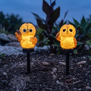 Könnte beinhalten: Zwei beleuchtete Gartenstecker in Eulenform mit warmgelbem Licht. Jede Eule hat große dunkle Augen und ein strukturiertes Federkleid und ist auf schwarzen Kunststoffstäben montiert. Die linke Eule hebt einen Flügel, die rechte hält einen Flügel am Kopf.