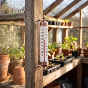 Pode incluir: Um termómetro de parede vertical com uma estrutura de metal cor de cobre e um tubo de vidro. A escala apresenta Fahrenheit e Celsius, com uma coluna de líquido vermelho a indicar a temperatura. Está montado num poste de madeira.