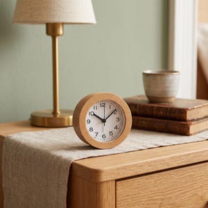 May include: A round analog alarm clock with a light-colored wooden frame, a white face, black numerals, and black hour, minute, and second hands. The clock features a simple circular design with a clear glass cover over the clock face.
