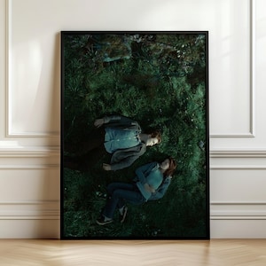 May include: A framed vertical wall art print featuring a high-angle view of two individuals lying on a patch of dark green grass. One individual wears a light blue button-down shirt and dark trousers, while the other wears a teal top and blue jeans.