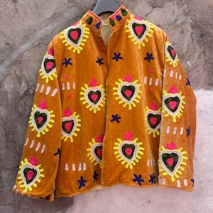 May include: A mustard yellow velvet jacket featuring embroidered heart motifs in bright pink, green, and yellow. The jacket has a high collar, long sleeves, and scattered dark blue star-shaped embroidery throughout the front and sleeve panels.