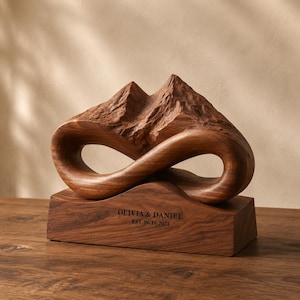 May include: A carved wooden sculpture featuring a mountain peak design integrated into an infinity symbol shape. The base is a rectangular wooden block engraved with the text OLIVIA & DANIEL EST. 06.18.2022 in a dark, sans-serif font.