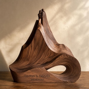 Op de afbeelding: Een donkerbruin houten sculptuur met twee abstracte figuren die elkaar omhelzen op de top van een bergachtige vorm. De basis is gegraveerd met de tekst OLIVIA & DANIEL en EST. 06.18.2022. De houtnerf is zichtbaar over het gladde, gebogen oppervlak.