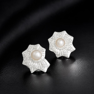 May include: A pair of stud earrings featuring a central round white pearl surrounded by a textured, star-shaped silver setting with intricate, granular detailing radiating from the center to the pointed edges of the metal frame.