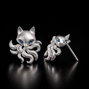 May include: A pair of silver-toned stud earrings shaped like a fox with multiple tails. Each fox features a textured, matte finish and two small, vibrant blue gemstone eyes. The earrings include a standard post backing for secure attachment.