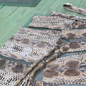 May include: Multiple rectangular and irregular pieces of natural snakeskin leather featuring a repeating diamond pattern in shades of tan, brown, cream, and grey. The textured reptile hide displays a distinct scale structure across all segments.