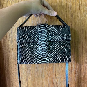 Puede incluir: Un bolso rectangular estructurado con un exterior de patrón de piel de serpiente en blanco y negro. El bolso incluye un asa superior, un cierre de solapa frontal con una lengüeta vertical y una correa de hombro negra, fina y ajustable.