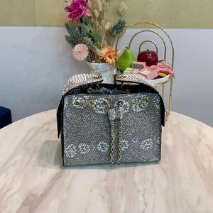 Puede incluir: Un bolso tipo cubo estructurado con un patrón de textura de piel de serpiente en gris y blanco, herrajes de metal dorado y un cierre de cordón con correas de cadena dobles. El bolso tiene una base rectangular y una abertura con ojales.