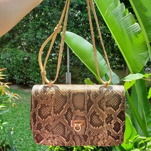 May include: A rectangular shoulder bag featuring a brown and black snakeskin pattern. The bag has a gold-tone metal clasp on the front flap and a thin, adjustable shoulder strap made of the same snakeskin material with gold-tone metal hardware.